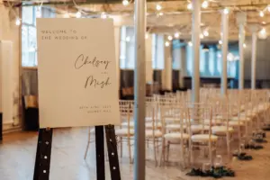 A wedding welcome sign on an easel reads Welcome to the wedding of Chelsey and Mish, 26th April 2025, at Holmes Mill in Clitheroe, set in a decorated indoor venue with rows of empty chairs and twinkling string lights.