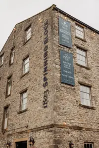 A stone building with large windows in Clitheroe displays signs reading 1823 Spinning Block and Holmes Mill, advertising a boutique hotel, spa, suites, bistro bar & grill, event functions, and weddings. The sky is overcast.