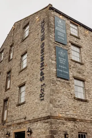 A stone building with large windows in Clitheroe displays signs reading 1823 Spinning Block and Holmes Mill, advertising a boutique hotel, spa, suites, bistro bar & grill, event functions, and weddings. The sky is overcast.