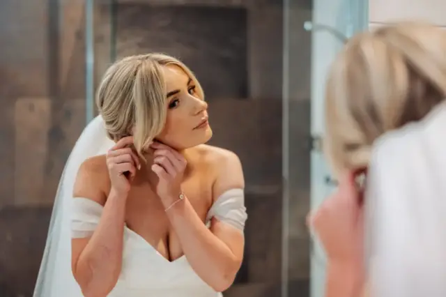 A bride with blonde hair in an off-the-shoulder white wedding dress adjusts her earring while looking in a mirror at Holmes Mill, Clitheroe, her reflection visible in the foreground.