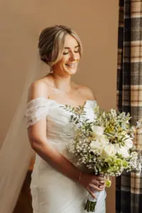 A smiling bride in an off-the-shoulder white wedding dress holds a bouquet of white flowers and greenery, standing near a window with plaid curtains at Holmes Mill, Clitheroe, on her special wedding day.