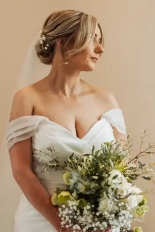 A bride in an off-the-shoulder white wedding dress holds a bouquet of white and green flowers. Her hair is styled in an updo with a veil and pearls, as she looks to the side at Holmes Mill in Clitheroe against a neutral background.