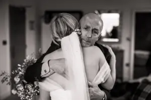 A man with a solemn expression hugs a woman in a wedding dress and veil from behind, holding her close in an emotional moment. The intimate scene unfolds indoors at Holmes Mill, Clitheroe, bathed in soft light on their wedding day.