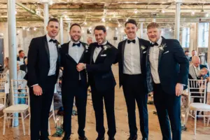 Five men in black tuxedos stand smiling together indoors at Holmes Mill, a rustic wedding venue in Clitheroe, with guests seated behind them and string lights hanging above.