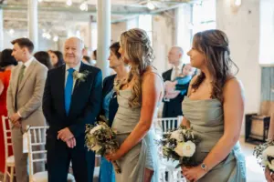 Two bridesmaids in light sage dresses hold bouquets among seated wedding guests at Holmes Mill. An older man in a suit and others look on in the bright, rustic Clitheroe wedding venue with white chairs and exposed beams.