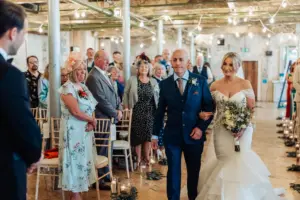 A bride in a white dress walks down the aisle at Holmes Mill, Clitheroe, with an older man in a blue suit, surrounded by guests at a rustic wedding venue glowing with string lights and candles.
