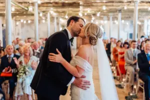 A bride and groom share a kiss at their Holmes Mill wedding ceremony in Clitheroe, surrounded by smiling, clapping guests. String lights hang from the rustic venue’s ceiling, creating a warm, festive atmosphere.