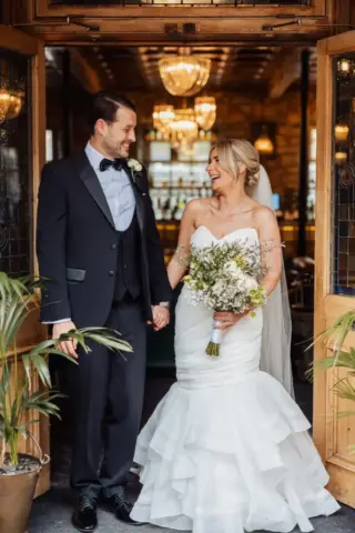 A bride in a strapless white gown holding a bouquet and a groom in a black tuxedo stand smiling at each other in the doorway of Holmes Mill, Clitheroe, with elegant lighting and plants around them, capturing the perfect wedding moment.