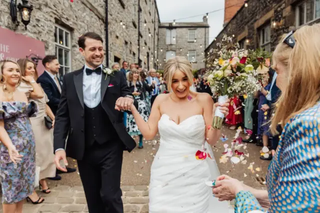 A bride in a white dress and a groom in a tuxedo smile joyfully as they walk outside at Holmes Mill in Clitheroe, holding hands, surrounded by guests throwing confetti. The festive wedding scene is filled with people in formal attire.