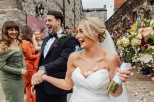 A bride and groom laugh joyfully while walking outside among guests after their Holmes Mill Wedding in Clitheroe. The bride holds a bouquet, flower petals on her dress, as guests smile and congratulate them in a festive atmosphere.