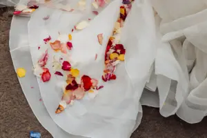 White sheer fabric, possibly a wedding dress, lies on the ground at Holmes Mill in Clitheroe, with scattered colorful flower petals in shades of red, pink, yellow, and orange on top of it.