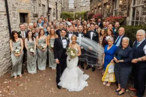 A large group of people pose outdoors for a wedding photo at Holmes Mill in Clitheroe, with the bride and groom smiling at the front. The guests, in formal attire, stand closely together amid stone buildings and scattered flower petals.