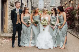 A bride in a white gown stands smiling with her four bridesmaids in matching sage green dresses and a groomsman in a suit; they hold bouquets outdoors on a stone path next to the rustic Holmes Mill, Clitheroe wedding venue.