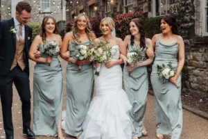 A smiling bride in a white dress walks arm-in-arm with four bridesmaids in light gray gowns and a man in a suit, all holding bouquets, on a path beside the stone buildings of Holmes Mill in Clitheroe, capturing a joyful wedding moment.