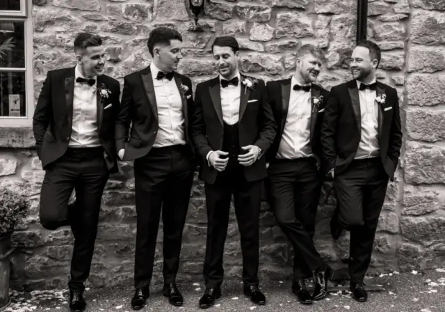 Five men in tuxedos stand and chat casually in front of a stone wall at Holmes Mill, Clitheroe, smiling and laughing together. They appear relaxed and are likely at a formal event, such as a wedding. The photo is in black and white.