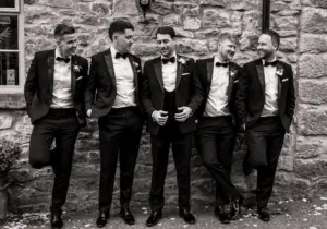 Five men in tuxedos stand and chat casually in front of a stone wall at Holmes Mill, Clitheroe, smiling and laughing together. They appear relaxed and are likely at a formal event, such as a wedding. The photo is in black and white.