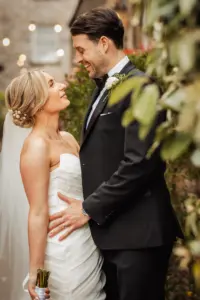 A bride and groom stand close together outdoors at Holmes Mill in Clitheroe, smiling lovingly at each other on their wedding day. The bride wears a strapless white gown and veil, holding a bouquet; the groom dons a black suit with a white boutonniere.