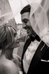 A bride and groom stand close together under a veil, smiling at each other with joy. The black-and-white photo at Holmes Mill captures their loving gaze and wedding attire, with blurred lights in the background.