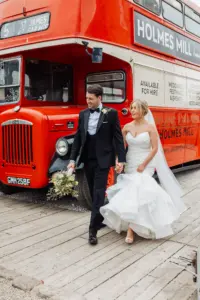 A bride and groom walk hand in hand in front of a vintage red double-decker bus with Holmes Mill signage in Clitheroe. The groom wears a black tuxedo; the bride, in a white strapless gown, holds her train, capturing the charm of their wedding day taken by a yorkshire wedding photographer