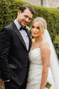A smiling bride in a white strapless dress and veil stands next to her groom in a black tuxedo at their Holmes Mill wedding. They pose closely together outdoors with lush Clitheroe greenery in the background.