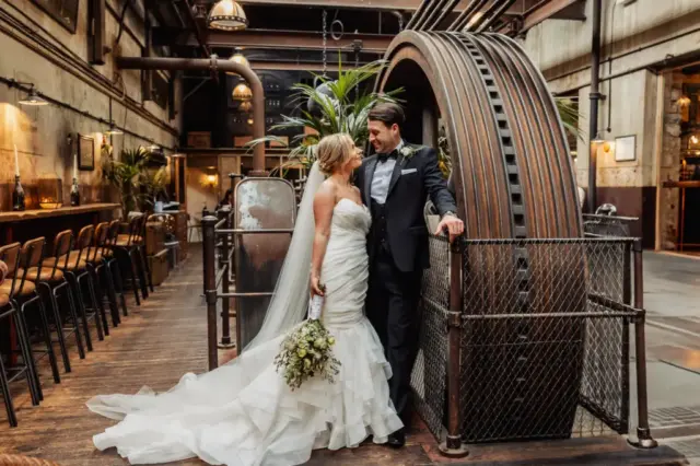 A bride and groom stand closely together, smiling, inside Holmes Mill in Clitheroe—a unique industrial-style wedding venue with exposed pipes, large gears, and wooden floors. The bride holds a bouquet and wears a white dress; the groom wears a black suit.