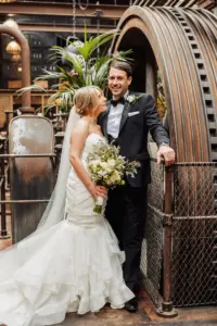 A bride in a white dress and veil holds a bouquet, smiling at the groom in a tuxedo. They stand together by industrial machinery at Holmes Mill in Clitheroe, with metal pipes and greenery in the background, celebrating their wedding day.