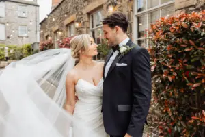 A bride in a white gown and veil stands arm-in-arm with her groom in a black tuxedo, smiling at each other outdoors by a stone building at Holmes Mill, Clitheroe, surrounded by greenery and red-leaved bushes on their wedding day.