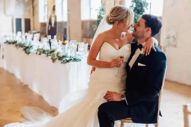 A bride in a white dress sits on the lap of her groom in a black suit at their Holmes Mill wedding in Clitheroe, both smiling and gazing at each other affectionately, with a beautifully decorated table in the background.