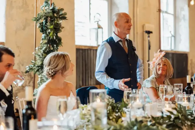 At Holmes Mill in Clitheroe, a man in a vest gives a heartfelt wedding speech as the bride and guests sit at a decorated table, smiling amid candles, flowers, and wine glasses.