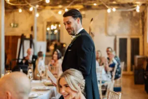 A man in a tuxedo stands giving a wedding speech at Holmes Mill in Clitheroe, holding a piece of paper. Guests sit at decorated tables, smiling under warm string lights in the rustic venue.