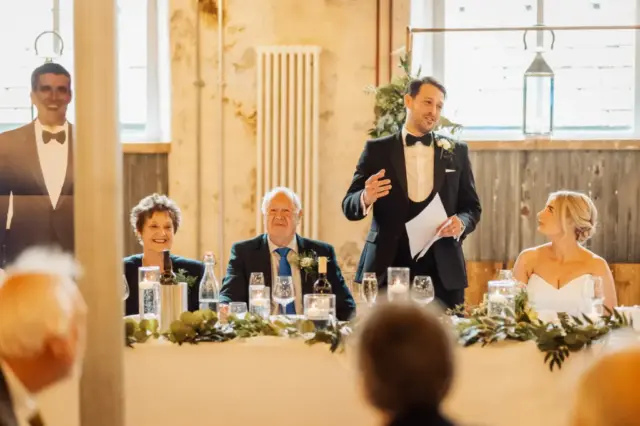 At a wedding reception at Holmes Mill in Clitheroe, a man in a tuxedo gives a speech behind a long table with seated guests, including an older couple and the bride. The warmly lit room features charming rustic decor.