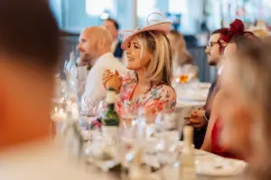 At a Clitheroe wedding at Holmes Mill, a woman in a pink floral dress and decorative hat smiles and claps at a formal event, seated at a table with glasses and bottles, surrounded by other well-dressed guests.