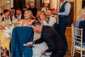 A bride smiles and looks surprised while sitting at a table as a man kneels beside her holding a microphone. Guests laugh and clap during a lively wedding reception at Holmes Mill in Clitheroe.