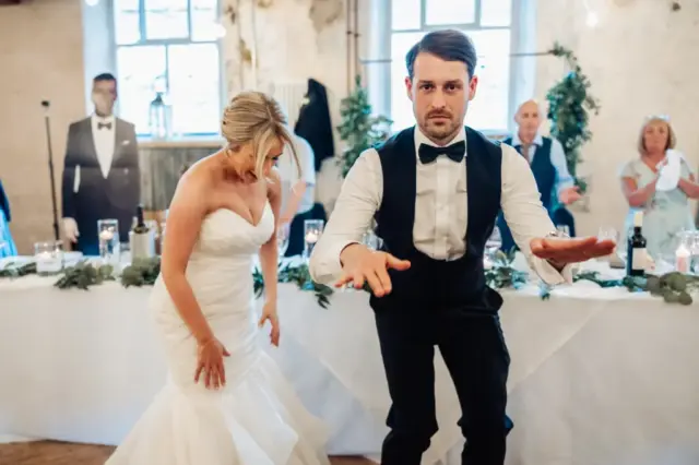 A bride in a white wedding dress and groom in a black vest and bow tie dance energetically at their Holmes Mill wedding reception in Clitheroe, while guests seated at a long table watch the lively moment unfold.