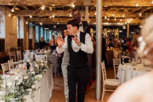 A man in formal attire walks through a decorated wedding reception hall at Holmes Mill in Clitheroe, gesturing with his hands, while a woman follows behind. Tables are set with candles, glasses, and greenery under string lights.