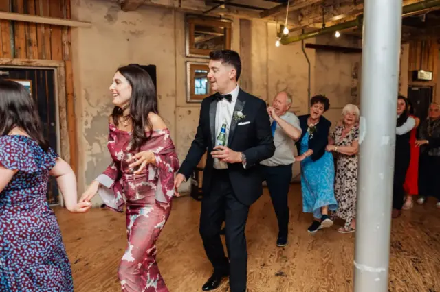 A group of people, led by a woman in a floral dress and a man in a suit holding a drink, dance in a conga line at a lively wedding reception inside Clitheroes rustic Holmes Mill, with wooden floors and exposed pipes.