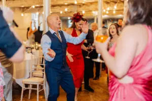 At a vibrant wedding at Holmes Mill in Clitheroe, a man in a blue suit enthusiastically dances beneath string lights, surrounded by elegantly dressed guests enjoying the celebration.