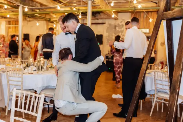 At a lively Holmes Mill wedding in Clitheroe, a man in a beige suit playfully hugs the legs of a man in black while other formally dressed guests mingle and dance in the warmly lit rustic venue.