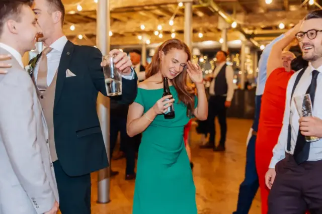 A group of people dressed in formal attire smile and laugh while holding drinks at a wedding in Holmes Mill, Clitheroe. String lights glow in the background as a cheerful woman in a green dress stands at the center of the lively indoor event.