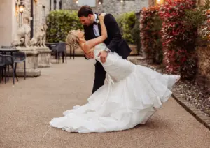 A groom in a black tuxedo dips his bride, who is wearing a strapless white wedding dress, as they kiss outdoors on a stone path lined with greenery and lights at their beautiful Clitheroe wedding.