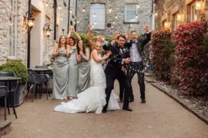 A bride and groom celebrate their Holmes Mill wedding outdoors in Clitheroe; the groom pops a champagne bottle, spraying everyone. Bridesmaids in light gray dresses and groomsmen in black suits cheer under string lights by stone walls.