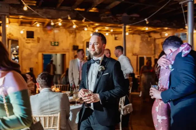 A man in a suit and bow tie, holding a drink, walks through the warmly lit wedding reception at Holmes Mill in Clitheroe. Guests are seated and mingling beneath string lights, surrounded by charming rustic decor.