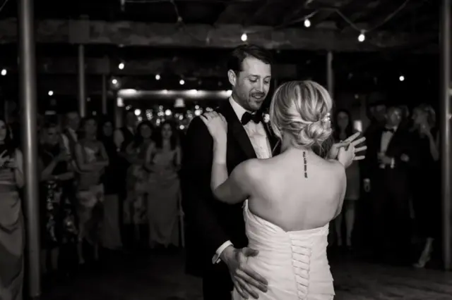 A bride and groom dance closely at their wedding reception in Holmes Mill, Clitheroe. The bride’s tattooed back faces the camera as guests watch and smile in the softly lit, rustic venue.
