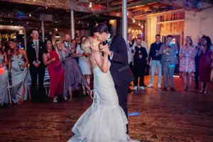 A bride and groom share a romantic first dance on a wooden floor as wedding guests watch and smile in a warmly lit, rustic Holmes Mill venue in Clitheroe.