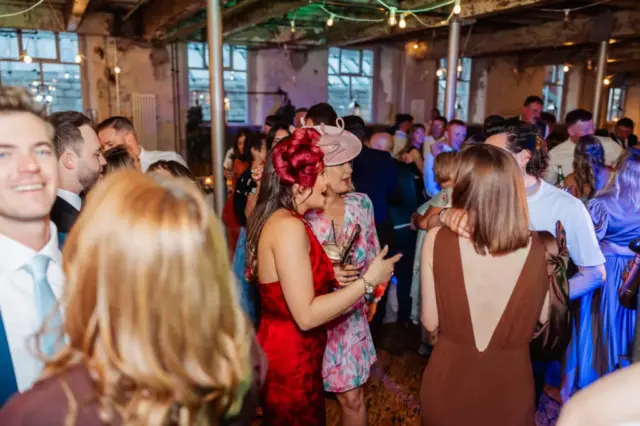 A lively indoor party at Holmes Mill in Clitheroe, with people dressed in formal attire mingling and chatting under string lights; some guests wear colorful dresses and fascinators, capturing the spirit of a stylish wedding celebration.