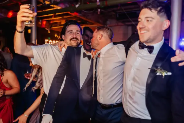 Three men in dress shirts and suits smile and pose together at a lively indoor wedding at Holmes Mill, Clitheroe. One holds a cardboard cutout in a suit, while another raises a drink for a selfie amid warm lighting and festive guests.