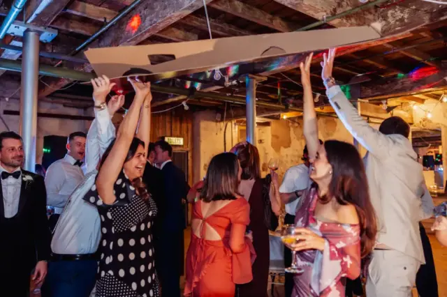 A group of people at a lively indoor party at Holmes Mill in Clitheroe, some holding a large cardboard cutout above their heads, smiling and celebrating a wedding under colorful lights and exposed wooden beams.