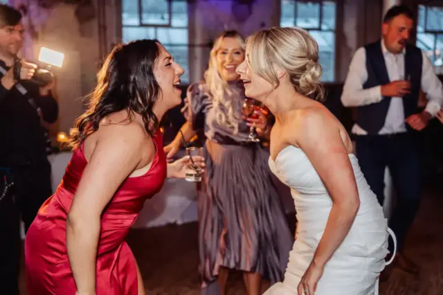Two women, one in a red dress and one in white, laugh and lean toward each other at a lively Holmes Mill wedding in Clitheroe, holding drinks as people dance and a photographer captures the joyful scene.