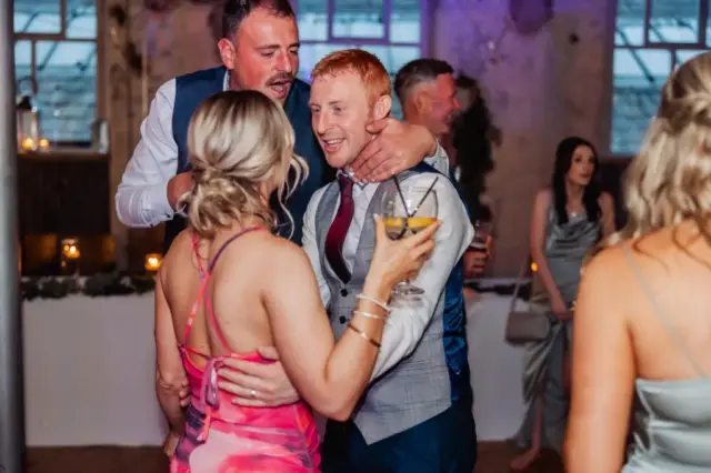 Three people are dancing and celebrating at an indoor wedding at Holmes Mill in Clitheroe. One man hugs another, who is smiling, while a woman in a pink dress holds a drink and faces them. Other guests and decorations are visible in the background.