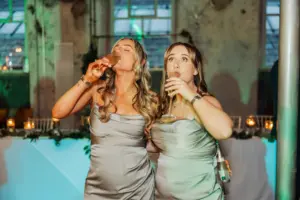 Two women in matching silver dresses enjoy wine at a Clitheroe wedding reception, possibly at Holmes Mill. Candles and greenery decorate the table behind them as they share a joyful moment.